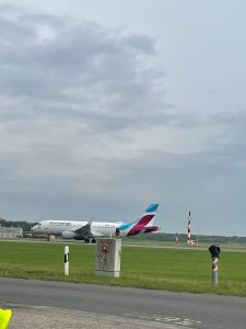 Ein Flugzeug mit buntem Heck auf einer Startbahn, umgeben von grünem Gras und unter einem bewölkten Himmel. In der Nähe stehen einige Verkehrsschilder und eine Person ist am Rand des Bildes sichtbar.