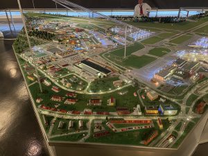 Modell einer Stadt oder eines Geländes mit verschiedenen Gebäuden, Straßen und Landschaftselementen, beleuchtet von kleinen Lichtern. Im Hintergrund sind weitere Details und eine Person sichtbar, die möglicherweise eine Führung durch das Modell anbietet.