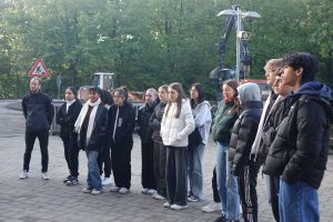Gruppe von Jugendlichen und jungen Erwachsenen steht in einer Reihe im Freien, umgeben von Bäumen. Sie tragen lässige Kleidung, darunter Jacken und Hoodies. Im Hintergrund ist ein Traktor und ein Verkehrsschild sichtbar. Die Gruppe wirkt aufmerksam und konzentriert.
