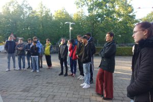 Gruppe von Jugendlichen und Erwachsenen steht im Freien, umgeben von Bäumen, während sie aufmerksam in eine Richtung schauen. Einige tragen Jacken und Sweatshirts, und die Stimmung wirkt konzentriert. Der Boden ist mit Pflastersteinen bedeckt.