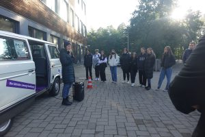 Eine Gruppe von Jugendlichen steht auf einem gepflasterten Platz, während eine Person in warmer Kleidung vor ihnen spricht. Neben der Gruppe steht ein weißer VW-Bus. Im Hintergrund sind moderne Gebäude und Bäume zu sehen. Das Bild vermittelt eine informative oder schulische Atmosphäre.