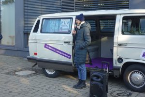 Eine Person steht neben einem weißen Lieferwagen, der teilweise offen ist. Die Person trägt eine dunkelgraue Jacke, eine Mütze und spricht in ein Mikrofon. Vor dem Fahrzeug steht ein Lautsprecher. Im Hintergrund sind moderne Gebäude und Fenster zu sehen. Der Boden ist gepflastert.