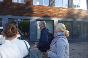 Drei Personen gehen auf einem Gehweg entlang eines modernen Gebäudes mit großen Fenstern. Die erste Person hat einen Dutt und trägt eine helle Jacke, die zweite Person ist männlich und trägt einen Rucksack, während die dritte Person eine helle Jacke und einen Schal trägt. Das Gebäude hat eine Holzverkleidung und ist von Tageslicht beleuchtet.
