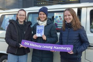 Drei Personen stehen vor einem weißen Lieferwagen und halten ein Banner mit der Aufschrift „Die kleinste Lesung Hamburgs“. Eine der Personen hält ein Buch in der Hand. Im Hintergrund sind Plakate zu sehen. Alle Personen tragen warme Winterkleidung.