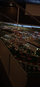 Modell einer Stadtlandschaft bei Nacht, beleuchtet mit zahlreichen kleinen Lichtern, die Straßen, Gebäude und Fahrzeuge darstellen. Der Blickwinkel zeigt die Modelle von oben, mit klaren Details der Infrastruktur und einer lebhaften Darstellung der Umgebung.