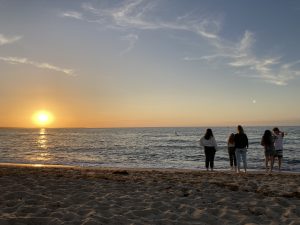 Silhouette von fünf Personen, die am Strand stehen und den Sonnenuntergang über dem Meer beobachten. Der Himmel ist sanft gefärbt, und die Wellen brechen sanft am Ufer.