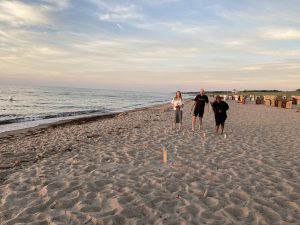 Drei Personen gehen am Strand entlang, der Sand ist hell und das Wasser ist ruhig. Im Hintergrund sind Strandkörbe zu sehen, und der Himmel hat eine sanfte Farbgebung kurz vor Sonnenuntergang.