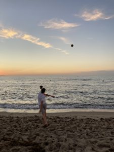 Eine Person steht am Strand und wirft einen Ball ins Meer. Im Hintergrund ist der Sonnenuntergang zu sehen, während sanfte Wellen an den Strand rollen. Der Himmel ist in sanften Farben gefärbt.