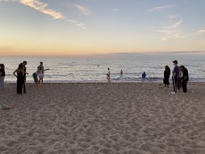 Menschen verbringen Zeit am Strand, einige stehen im Sand, während andere im Wasser spielen. Der Himmel ist in sanften Farben der Dämmerung gefärbt.