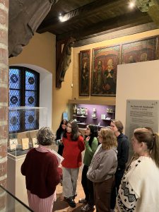 Eine Gruppe von sechs Personen hört einer Führerin in einem Museum aufmerksam zu. Im Hintergrund sind historische Gemälde und Ausstellungsstücke zu sehen. Das Licht ist sanft und die Wände sind in warmen Farben gehalten. Die Personen sind unterschiedlich gekleidet und stehen um einen Ausstellungsbereich herum.