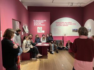 Eine Gruppe von Menschen sitzt in einem Museum auf einer Bank und hört einer Referentin zu, die vor ihnen steht. Die Wände sind in einem dunklen Rosaton gestrichen, und es gibt informative Texte an den Wänden. Die Atmosphäre ist freundlich und informativ, mit Fokus auf das Lernen und den Austausch von Ideen.