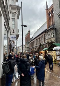 Menschenmenge auf einer Stadtstraße, umgeben von historischen Gebäuden und Geschäften. Der Himmel ist bewölkt, und es scheint, als ob es regnet. Stände mit Produkten sind im Hintergrund sichtbar.