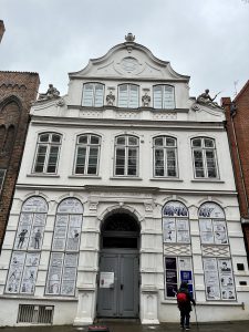 Historisches Gebäude mit weißer Fassade und mehreren Fenstern, das mit Plakaten und Bildern geschmückt ist. Der Eingang ist zentral angeordnet und von zwei hohen Fenstern flankiert. Über dem Eingang befindet sich ein geschwungenes Giebelfeld. Der Himmel ist bewölkt. Im Vordergrund ist eine Person zu sehen, die an dem Gebäude vorbeigeht.