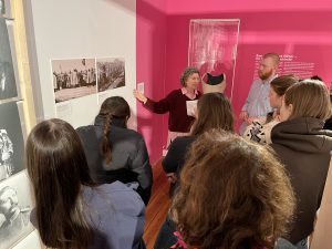 Eine Gruppe von Menschen hört einer älteren Frau zu, die in einem Ausstellungsraum mit pinken Wänden steht. Sie zeigt auf Bilder an der Wand, während ein Mann neben ihr steht. Im Hintergrund sind weitere Personen zu sehen, die auf die Präsentation achten.