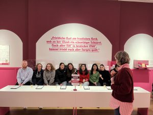 Eine Gruppe von sieben Personen sitzt an einem Tisch in einem Raum mit pinken Wänden. Vor ihnen befindet sich ein Tisch mit Notizen oder Dokumenten. Im Hintergrund hängt ein Zitat an der Wand in deutscher Sprache. Eine Frau steht am Tisch und spricht zu der Gruppe.