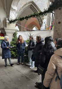 Eine Gruppe von sechs Personen steht in einem überdachten Bereich, umgeben von Weihnachtsbäumen. Eine Person in der Mitte liest aus einem Notizbuch vor. Die anderen hören aufmerksam zu. Es ist eine graue, winterliche Atmosphäre, und die Personen tragen warme Kleidung. Im Hintergrund sind architektonische Elemente und festliche Dekorationen sichtbar.
