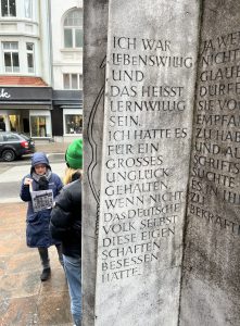Eine Person steht an einem monumentalen Stein, auf dem eine Inschrift zu lesen ist. Die Inschrift beginnt mit „Ich war lebenswürdig und das heißt lernwillig sein...“. Im Hintergrund ist ein weiteres Gebäude und eine weitere Person zu sehen, die ein Plakat oder eine Zeitung hält.