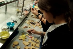 Eine Person dekoriert Plätzchen auf einem Backblech. Vor ihr sind verschiedene Plätzchen in unterschiedlichen Formen, die mit buntem Zuckerguss und Streuseln verziert werden. Es gibt mehrere Schalen mit Zuckerguss in verschiedenen Farben. Die Umgebung wirkt hell und freundlich.