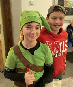 Zwei Jungen stehen lächelnd zusammen. Der Junge links trägt ein grünes Kostüm und eine grüne Mütze, die an eine Figur aus einem Videospiel erinnert. Er hält einen roten Lollipop in der Hand. Der Junge rechts trägt ein rotes Sweatshirt mit der Aufschrift 