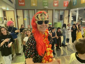 Eine fröhliche Person mit rotem Haar, Sonnenbrille und Blumenkranz steht im Vordergrund. Im Hintergrund sind viele Kinder und Jugendliche in bunten Kostümen, die an einer Feier teilnehmen. Die Umgebung ist hell erleuchtet und dekoriert.