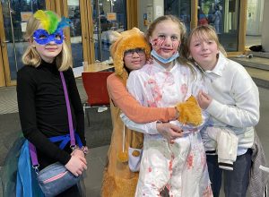 Vier Kinder in kostümierten Outfits stehen zusammen. Ein Mädchen trägt eine bunte Maske und ein schwarzes Oberteil, ein anderes ist als Löwe verkleidet, während ein drittes Kind in einem blutigen weißen Kostüm mit Mundschutz zu sehen ist. Das vierte Kind trägt einen weißen Pullover. Sie lächeln und posieren in einer hellen Innenumgebung.