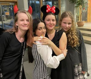 Vier junge Mädchen posieren lächelnd zusammen in einem Innenraum. Ein Mädchen trägt ein schwarzes Oberteil und hat rote Teufelshörner, ein anderes trägt ein Outfit mit Minnie-Maus-Ohren, während die beiden anderen in modischer Kleidung gekleidet sind. Sie stehen eng beieinander und wirken fröhlich. Im Hintergrund sind Pflanzen und ein moderner Eingangsbereich sichtbar.