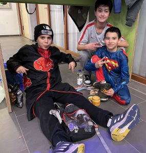 Drei Jungen sitzen in einem Raum. Der Junge links trägt ein schwarzes Outfit mit roten Details und eine Mütze. Der Junge in der Mitte trägt ein graues T-Shirt und lächelt. Der Junge rechts trägt ein Superhelden-Kostüm. Auf dem Boden vor ihnen liegen einige Snacks und Getränke. Die Umgebung wirkt freundlich und ungezwungen.
