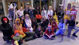Gruppenszene von Kindern und Jugendlichen in bunten Kostümen, die in einer Halle posieren. Einige tragen Tierkostüme oder fantasievolle Outfits, während andere sich als bekannte Charaktere verkleidet haben. Die Gruppe steht sowohl stehend als auch sitzend zusammen und lächelt in die Kamera.