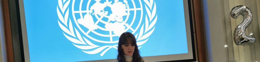 Eine Person steht vor einer Präsentation mit dem Logo der Vereinten Nationen auf einem blauen Hintergrund.