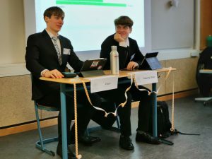 Zwei junge Männer in Anzügen sitzen an einem Tisch, der mit einer Wasserflasche und Laptops ausgestattet ist. Sie scheinen an einer Präsentation oder Diskussion teilzunehmen. Im Hintergrund ist eine Präsentationsfolie sichtbar.