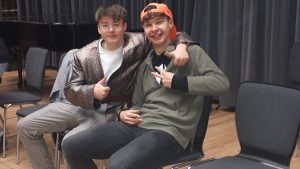 Zwei junge Männer sitzen in einem Raum auf Stühlen. Der erste trägt eine schimmernde Jacke und gibt einen Daumen hoch, während der zweite in einem grauen Hoodie mit einer orangefarbenen Mütze sitzt und ein Peace-Zeichen zeigt. Im Hintergrund sind dunkle Vorhänge und Stühle sichtbar.