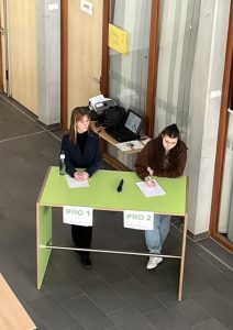 Zwei Personen sitzen an einem grünen Tisch, der mit den Schildern 