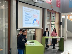 Eine Veranstaltungsszene in einem modernen Raum mit einem großen Bildschirm im Hintergrund, auf dem Informationen präsentiert werden. Im Vordergrund stehen drei Personen an grünen Tischen. Eine Person hält ein Mikrofon und spricht. Die anderen beiden Personen hören zu und sind in ein Gespräch vertieft. Im Raum sind bunte Flaggen und eine helle, offene Atmosphäre.