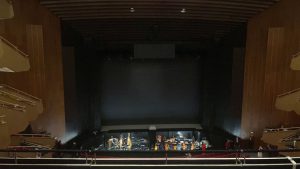 Blick auf eine leere Bühne in einem Theater mit einer dunklen Hintergrundwand. Die Zuschauerbereiche sind sichtbar, mit Holzverkleidung und Stufen. Die Beleuchtung ist gedämpft, und es sind keine Personen auf der Bühne oder im Publikum zu sehen.