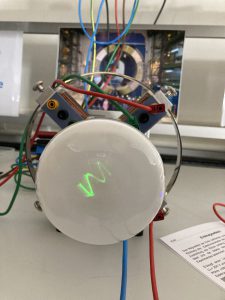 Eine technische Vorrichtung mit einem runden, weißen Gehäuse in der Mitte, umgeben von Metallringen und bunten Drähten. Auf dem Gehäuse ist eine grüne, leuchtende Welle sichtbar. Im Hintergrund ist ein weiteres Element zu sehen, das die Struktur ergänzt. Rechts liegt ein Zettel mit Informationen.