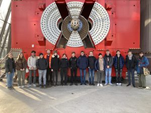 Gruppe von Personen steht vor einer großen, roten Maschine mit einem kreisförmigen Design und großen Schrauben. Die Personen sind unterschiedlich gekleidet und lächeln, während sie für das Foto posieren. Im Hintergrund ist eine industrielle Umgebung sichtbar.