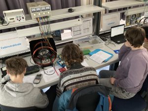 Drei Personen sitzen an einem Tisch mit technischen Geräten und Laptops. Die Umgebung ist ein Labor oder eine Werkstatt, mit Kabeln und Messgeräten auf dem Tisch. Die Personen sind in konzentrierte Arbeit vertieft, während sie Unterlagen und Geräte nutzen.