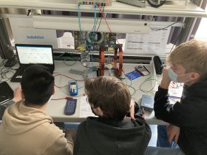 Drei Personen sitzen an einem Tisch und betrachten einen Laptop-Bildschirm. Auf dem Tisch befinden sich verschiedene Geräte und Kabel, darunter ein Modell oder eine Maschine, die mit dem Laptop verbunden ist. Eine Person trägt eine Maske. Der Bildschirm zeigt eine grafische Darstellung an.