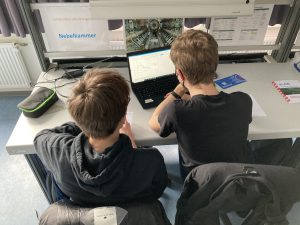 Zwei Jugendliche sitzen an einem Tisch und schauen auf einen Laptop, der geöffnet ist. Sie sind von hinten zu sehen. Auf dem Tisch liegen ein paar Gegenstände, darunter eine schwarze Tasche und ein Dokument. Im Hintergrund sind einige Plakate oder Informationen sichtbar. Die Szene ist hell beleuchtet.