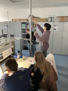 Schüler arbeiten in einem Labor. Ein Schüler steht an einem Tisch und justiert ein Gerät, während zwei weitere Schüler am Tisch sitzen und Notizen machen. Im Hintergrund sind Regale mit Materialien und Dokumenten sichtbar.