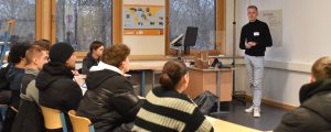 Ein Referent steht vor einer Gruppe von Schülern in einem Klassenraum. Die Schüler sitzen an Tischen und hören aufmerksam zu. Im Hintergrund sind Fenster mit Tageslicht und ein Bildschirm zu sehen.