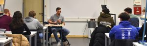 Ein Dozent sitzt an einem Tisch und präsentiert vor einer Gruppe von Studierenden, die ihm gegenüber sitzen. Im Hintergrund sind eine Tafel und einige technische Geräte zu sehen. Die Studierenden sind in verschiedenen Kleidungsstücken gekleidet, einige haben Kapuzenpullover an.