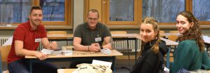 Vier Personen sitzen an einem Tisch in einem Raum mit großen Fenstern. Sie schauen freundlich in die Kamera. Zwei Männer und zwei Frauen sind sichtbar, alle tragen Namensschilder. Auf dem Tisch liegen einige Unterlagen. Der Raum wirkt hell und einladend.