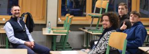 Vier Personen sitzen in einem Klassenzimmer. Im Vordergrund lächelt ein Mann mit Bart, der einen dunklen Weste trägt. Neben ihm sitzen drei jüngere Männer, von denen einer Daumen hoch zeigt. Im Hintergrund sind leere Tische und Stühle zu sehen, die auf den Tischen gestapelt sind, sowie Fenster, durch die Tageslicht fällt.