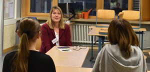 Eine Frau mit langen, blonden Haaren sitzt an einem Tisch und spricht mit zwei Personen, die ihr gegenüber sitzen. Die Szene spielt sich in einem Klassenraum ab, der mit Tischen und Stühlen ausgestattet ist. Die Frauen, die ihr gegenüber sitzen, sind von hinten zu sehen. Die Atmosphäre wirkt freundlich und kommunikativ.