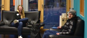 Zwei Personen sitzen in einem modernen Wartebereich. Eine Frau mit langen Haaren lächelt, während sie auf einem schwarzen Sofa sitzt. Ihr rechter Fuß ruht auf dem linken Bein. Ein Mann in einer dunklen Jacke und einer Mütze sitzt schräg gegenüber. Im Hintergrund sind große Fenster, durch die man eine städtische Umgebung sehen kann. Die Wände sind in einem hellen Blau gestrichen.
