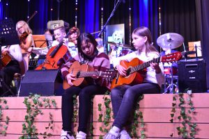 Zwei Mädchen sitzen auf einer Holzplattform und spielen Gitarre. Im Hintergrund sind weitere Kinder zu sehen, die verschiedene Musikinstrumente spielen, darunter Geigen und Schlagzeug. Die Bühne ist mit Lichtern und Pflanzen dekoriert.