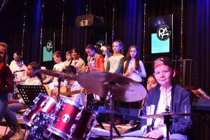 Eine Gruppe von Kindern und Jugendlichen steht auf einer Bühne, während ein Junge am Schlagzeug sitzt. Im Hintergrund sind weitere Musiker zu sehen, die verschiedene Instrumente spielen. Die Bühne ist beleuchtet, und im Hintergrund sind zwei Bildschirme mit dem Logo der Veranstaltung sichtbar. Die Atmosphäre wirkt lebhaft und musikalisch.