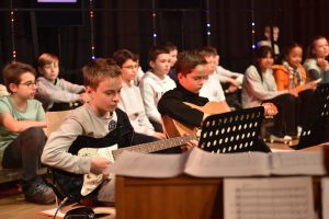 Gruppe von Kindern, die auf einer Bühne sitzen und Gitarre spielen. Einige Kinder halten Notenständer, während sie konzentriert musizieren. Im Hintergrund sind weitere Kinder zu sehen, die ebenfalls an der Aufführung teilnehmen. Die Bühne ist mit Lichtern dekoriert.