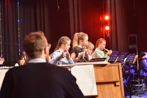Eine Gruppe von Musikern, darunter Kinder und Jugendliche, spielt Instrumente auf einer Bühne. Im Vordergrund ist ein Musiker zu sehen, der einen Blick auf die Notenblätter wirft. Im Hintergrund sind weitere Musiker, die konzentriert musizieren. Die Bühne ist mit farbigen Lichtern beleuchtet, die eine festliche Atmosphäre schaffen.
